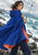 An adult woman in a windblown cobalt cape with coral lining holds a transparent umbrella beside a rocky, foaming sea.