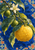 Golden citrus fruit, two green fruits, and a white blossom on a rain-beaded branch against cobalt blue tile-like motifs.