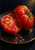 Split red heirloom tomato with glistening seed chambers beside an intact ribbed tomato on a dark plate, with loose seeds below.
