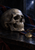 Human skull beside a folded blue and crimson sash and a round medal on a dark tabletop, with a moonlit opening behind.