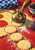 A brass-and-dark-metal rotary cutter presses into rolled dough, with scalloped rounds, scattered flour, checked cloth, and dark currants.