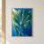 Large blue-green fan palm leaf cropped at the top and sides, with a smaller curled frond beside the stem against a vivid blue wall.; frame shown for styling only