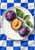 Three purple plums, including one cut open, sit on a white plate over blue-and-white checkerboard cloth with green leaves.