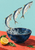 Three silver fish leaping above an indigo bowl with splashing water, a cut lemon, and leaves on a coral tabletop.