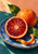 A cut blood orange sits before a whole orange on a blue plate, with green leaves and curling peel against a peach wall and teal tabletop.