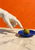 A pale sculpted-looking hand reaches toward an upright green olive on a cobalt-blue plate, with an orange wall behind it.