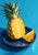 Halved pineapple with its leafy crown and a separate wedge on a dark blue plate against a turquoise background.