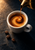 White espresso cup with a swirling crema spiral as coffee pours from a cropped brass kettle, with three beans on a dark counter.