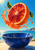 Cut blood-orange half floating above a deep cobalt bowl, surrounded by an arcing juice splash, droplets, and green leaves with sea and coast behind.