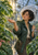 Curly-haired woman in a dark green jacket holding a clear umbrella and extending an open hand beneath a large wet monstera leaf in a sunlit glasshouse.