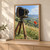 A large-format bellows camera on a wooden tripod stands beside a red poppy, with rolling green hills, distant buildings, and a pale-blue cloudy sky.; frame shown for styling only