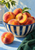 Blue-and-cream striped bowl filled with peaches, including a cut peach, beside a whole peach and green cloth.