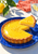 Glossy lemon tart on a cobalt plate with a slice lifted above it, beside a lemon, leaves, coral cloth, and a glass of water.