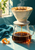 Ceramic pour-over dripper and glass carafe as coffee pours, with steam, teal linen, and orange peel in warm window light.