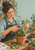 Woman in a pale blue-green shirt and dark apron trims a leafy plant above a terracotta pot with orange-handled pruning shears at a wooden workbench.