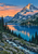 Snow-streaked mountain above a cobalt lake at sunset, with evergreen forest, a small red-roofed building, and a foreground footbridge.