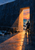 A cyclist and bicycle at a dark sea-wall opening, facing a sunset over the ocean as orange light reflects across wet stone.