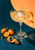 Cut-crystal coupe with amber drink, spherical ice, and curled orange peel, with kumquats and leaves against a teal and apricot background.