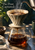 Ceramic pour-over brewer on a glass carafe as a kettle pours water into coffee grounds, with the title The Pour, Captured.