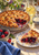 Golden lattice berry pie with a slice on a patterned plate, surrounded by berries, flowers, blue cloth, and a glass pitcher.
