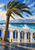 A wind-bent palm before a white seaside arcade with cobalt doors, two walkers, blue sea, and distant mountains.