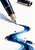 A dark fountain pen with gold-toned trim touches white paper beside a flowing blue-black ink stroke and separate cap.