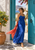A woman in a cobalt-blue dress holds a coral scarf between open turquoise doors, beside a potted citrus tree in a sunlit courtyard.