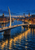 A modern cable-supported pedestrian bridge crosses a dark river between warmly lit buildings at blue hour.