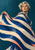 A short-haired woman lifts a sweeping cobalt-and-ivory striped cape against a deep teal background.