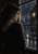 Dark-haired adult woman in three-quarter rear profile looking toward a warm-lit pane through a rain-streaked city window.