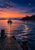 A small lit fishing boat crosses a twilight harbor toward a lighthouse as a crescent moon hangs above the water.