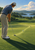 Golfer bending over a putter beside a white ball on a green, with a distant cup, lake, wooded hills, and clouds.