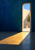 Tall opening in a deep-blue wall reveals a pale courtyard and small tree as a golden beam falls diagonally across the floor.