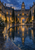 A symmetrical stone courtyard with a central clock tower, glowing windows, wet paving, and a reflecting pool at blue hour.
