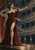 Horned woman in a long burgundy gown reaches toward an audience in a multi-tiered theater as pale bands sweep across the stage.