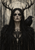 Adult dark-haired gothic witch with antler-like head adornments and a black raven on her shoulder against a distressed pale background.