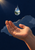 An open palm reaches toward a suspended clear water droplet containing tiny green hills, trees and sky against a midnight-blue background.