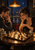 Two adults lean over a chessboard at a café table beneath a warm pendant lamp, with a moon and city lights in the indigo window.