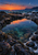 Rocky tidal pool reflecting an apricot sunset and crescent moon, with foamy edges, waves and a distant wooded headland.