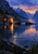 Warmly lit wooden boathouse and footbridge beside a mountain lake, with golden light reflected in rippling water and rocks and reeds at the shore.
