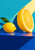 Upright cut lemon beside a smaller whole lemon with a green leaf on a cobalt-blue ledge against a turquoise wall and warm diagonal light.