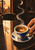 Blue-and-ivory espresso cup and saucer with swirling crema on a dark café table, held by a hand beneath curling steam and window light.