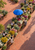 Overhead diagonal view of an open-air flower market with bouquet-filled stalls, vendors, terracotta paving, and a blue parasol.