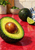 Textured still life of a cut avocado with a large brown pit, whole avocado, lime wedge, blue-striped towel, bowl, and plant on a red counter.