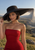 Fashion portrait of a woman in a red strapless dress holding a broad black hat in a rocky, grassy landscape.