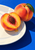 Cut peach with exposed pit and whole peach with a green leaf on a white plate against a cobalt-blue surface.