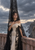 Veiled woman in an ivory gown beside a stone sea wall, with a stormy sea and distant cliffside buildings.