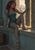 Gray-skinned, pointed-eared woman with curly red hair seated on a stone windowsill, reaching toward a brass wall fixture with a sunlit town and mountains beyond.