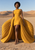 Barefoot short-haired woman in a golden sleeveless dress with expansive radiating pleats walking across a desert with dunes and cracked ground.