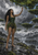 Barefoot woman in leafy green attire reaching toward moss-covered branches beside a lake under a lightning storm.
